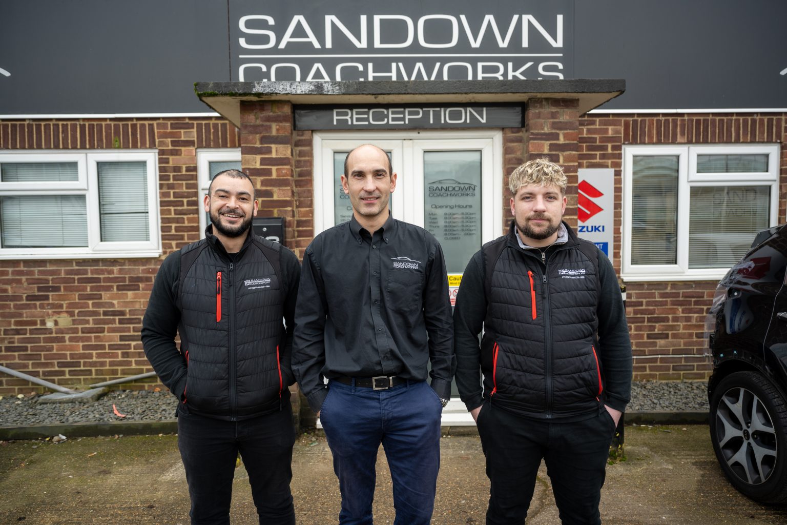 Three Sandown Coachworks team members in branded uniforms standing outside the reception entrance, with Suzuki manufacturer signage visible on the building