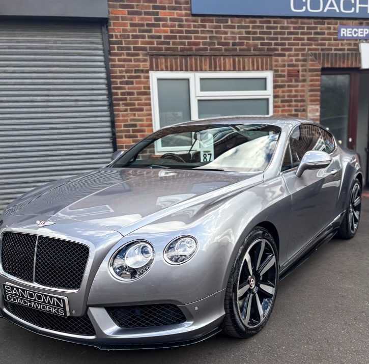 A silver Bentley Continental GT coupe parked on the tarmac outside a brick building featuring a Sandown Coachworks reception sign.