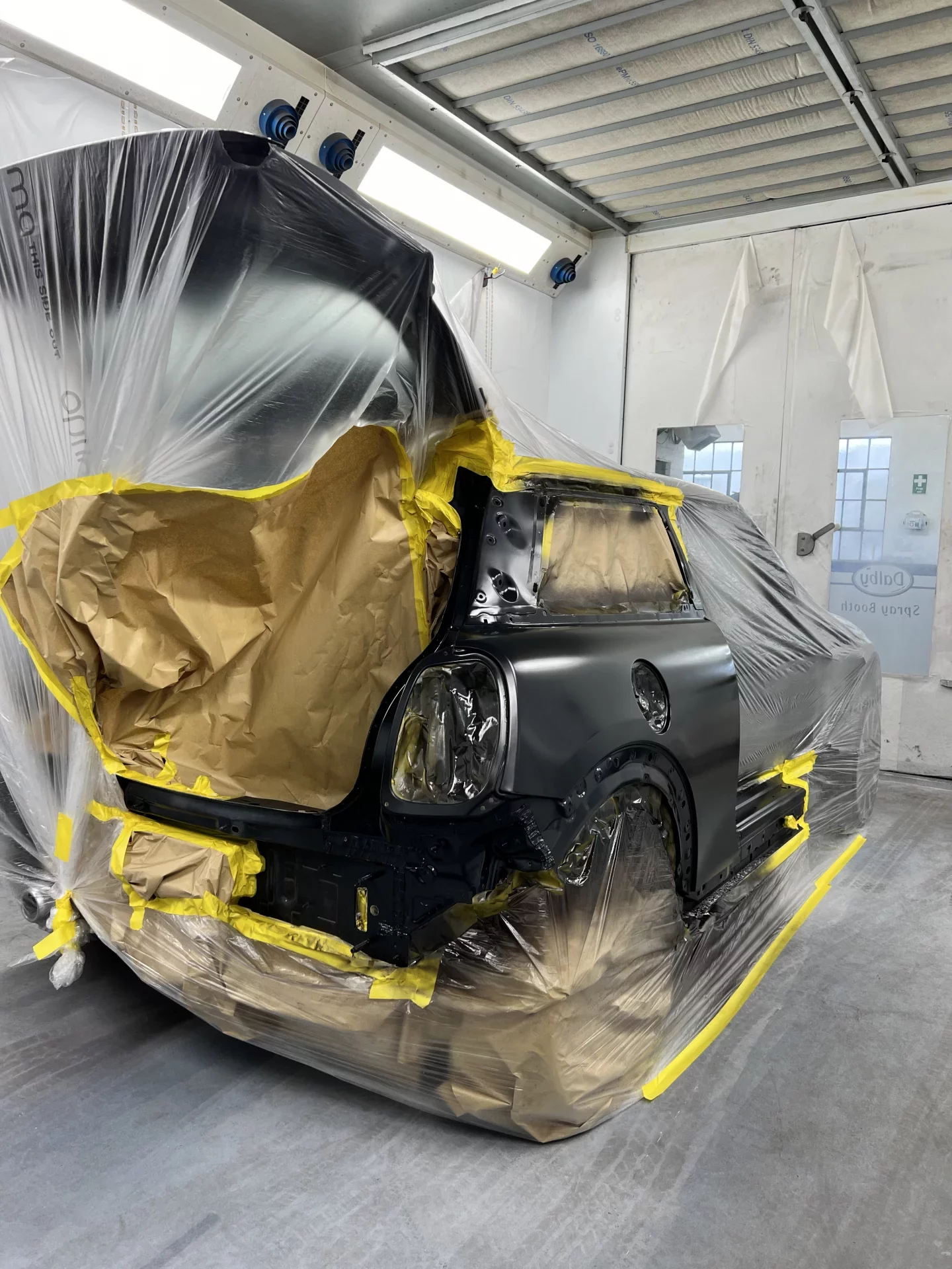 A Mini Cooper masked with protective film and paper inside a professional spray booth, undergoing a factory-standard paint application.