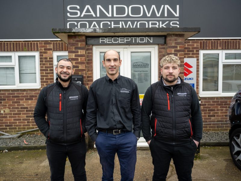 Three Sandown Coachworks team members in branded uniforms standing outside the reception entrance, with Suzuki manufacturer signage visible on the building