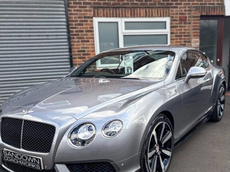 A silver Bentley Continental GT coupe parked on the tarmac outside a brick building featuring a Sandown Coachworks reception sign.