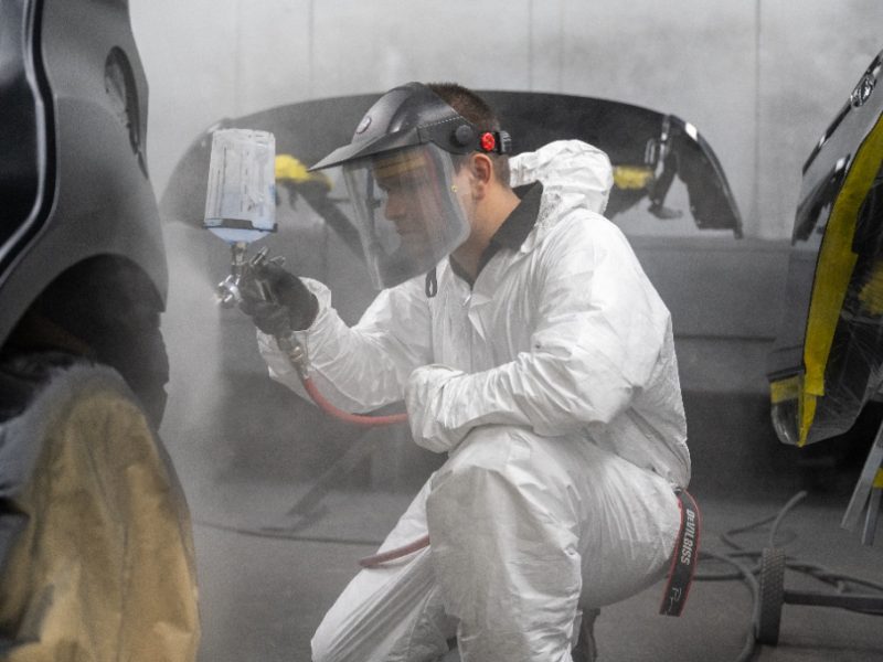 A technician wearing a white protective hazmat suit, face shield, and respirator mask uses a professional spray gun to paint a car panel inside a controlled spray booth.