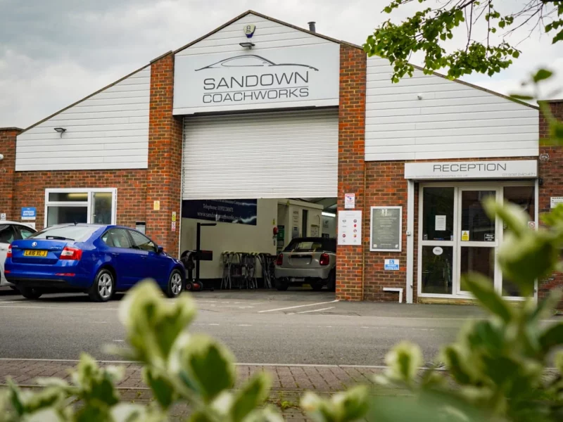 The exterior of Sandown Coachworks in Shepperton, showing the main workshop entrance, reception area, and various manufacturer approval signs on the brick wall.