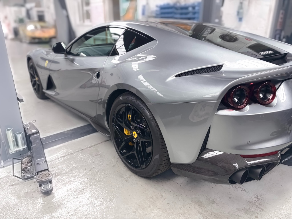 Rear three-quarter view of a metallic silver-grey Ferrari 812 Superfast inside a clinical workshop bay, highlighting complex body lines, quad exhaust tips, and deep gloss reflection.