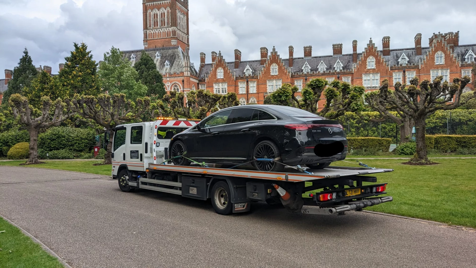 A black Mercedes-Benz EQS being transported on a Sandown Coachworks recovery truck, featuring the Holloway College building in the background.
