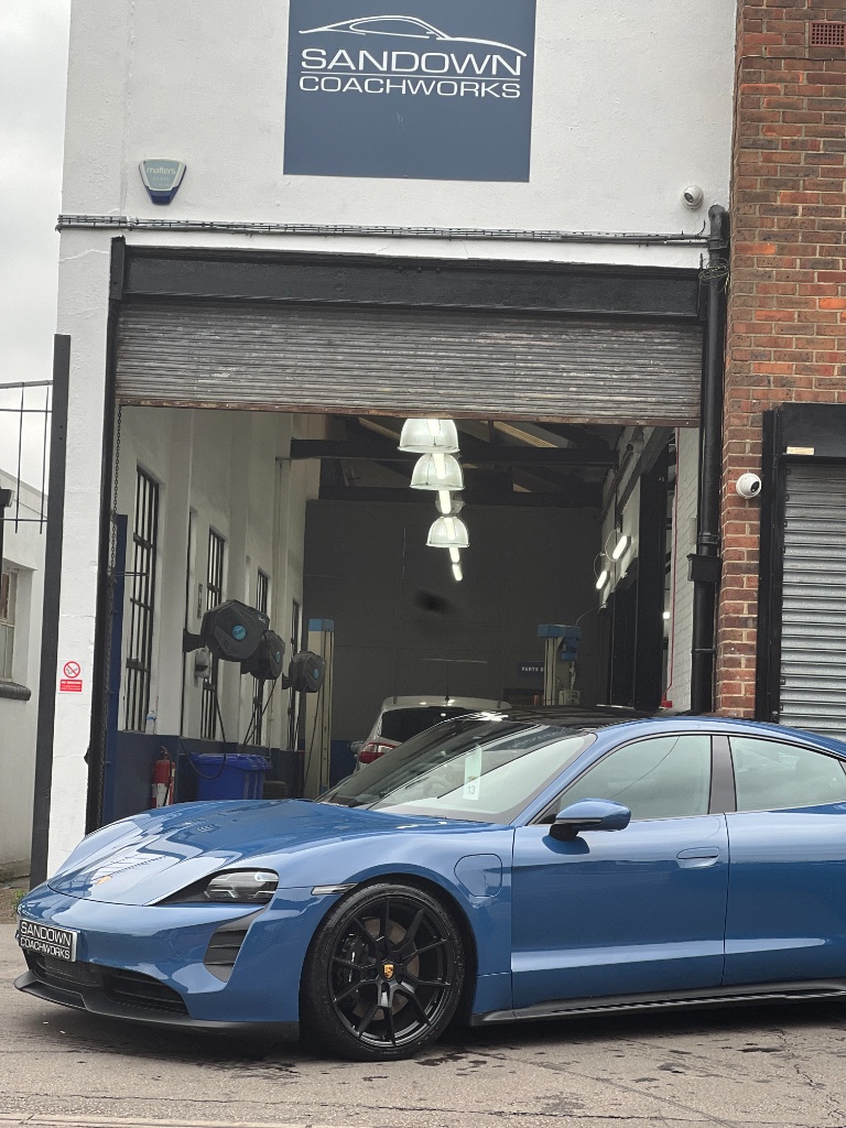 A metallic blue Porsche Taycan parked in front of an open workshop bay at Sandown Coachworks, with the garage sign visible above the entrance