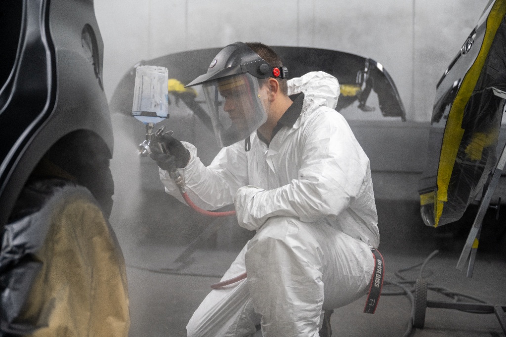 A technician wearing a white protective hazmat suit, face shield, and respirator mask uses a professional spray gun to paint a car panel inside a controlled spray booth.