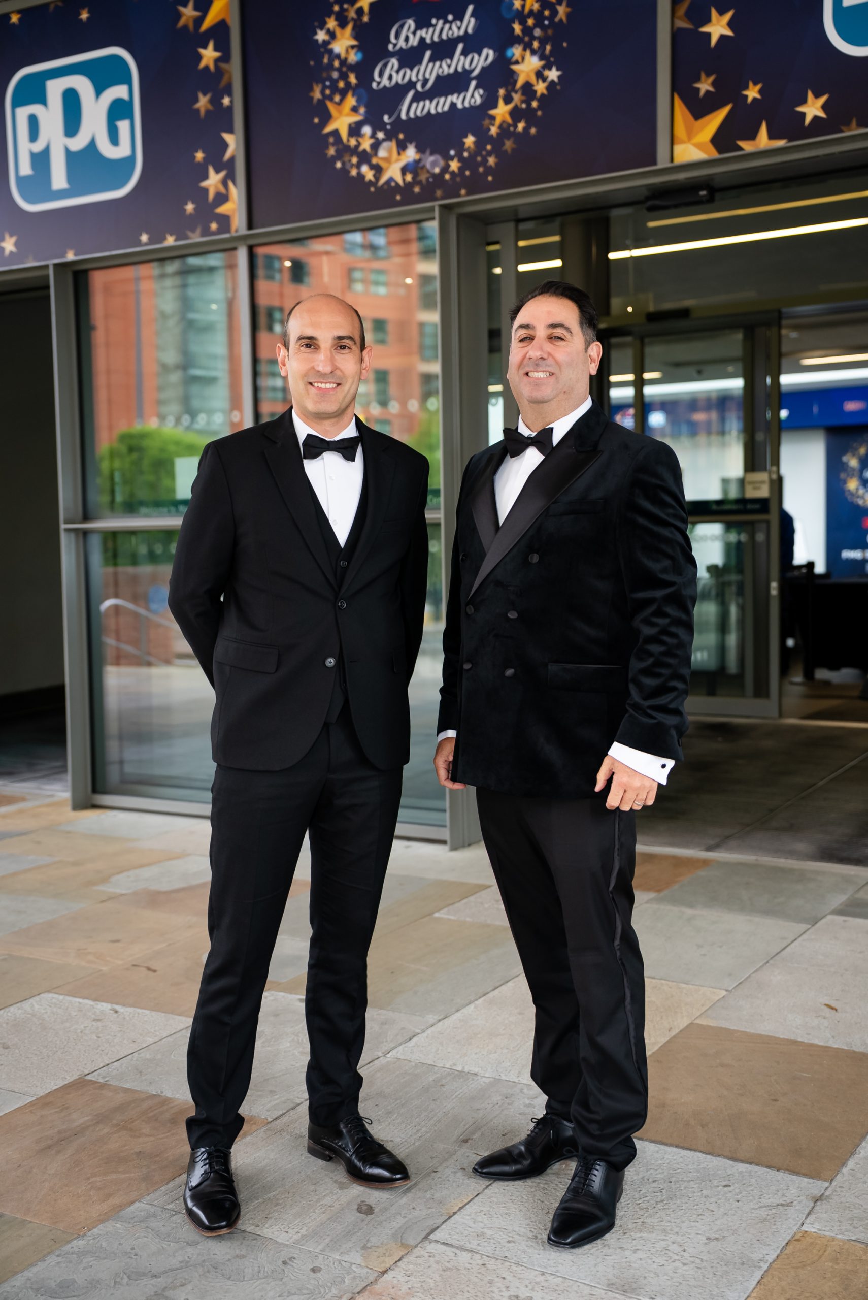 Two representatives from Sandown Coachworks wearing black tie attire standing in front of the British Bodyshop Awards entrance.