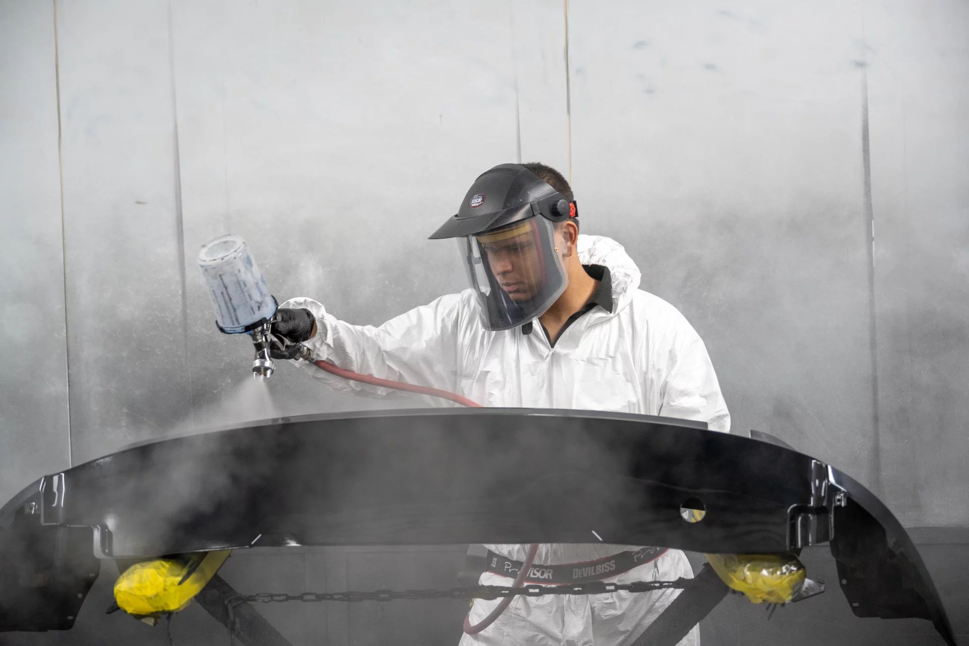 A highly skilled Sandown Coachworks technician in a protective suit and respirator mask expertly applying paint to a car bumper in a professional spray booth.