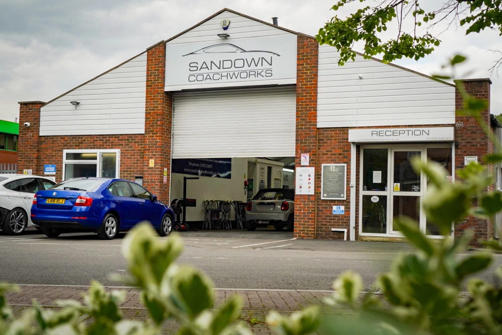 The exterior of Sandown Coachworks in Shepperton, showing the main workshop entrance, reception area, and various manufacturer approval signs on the brick wall.