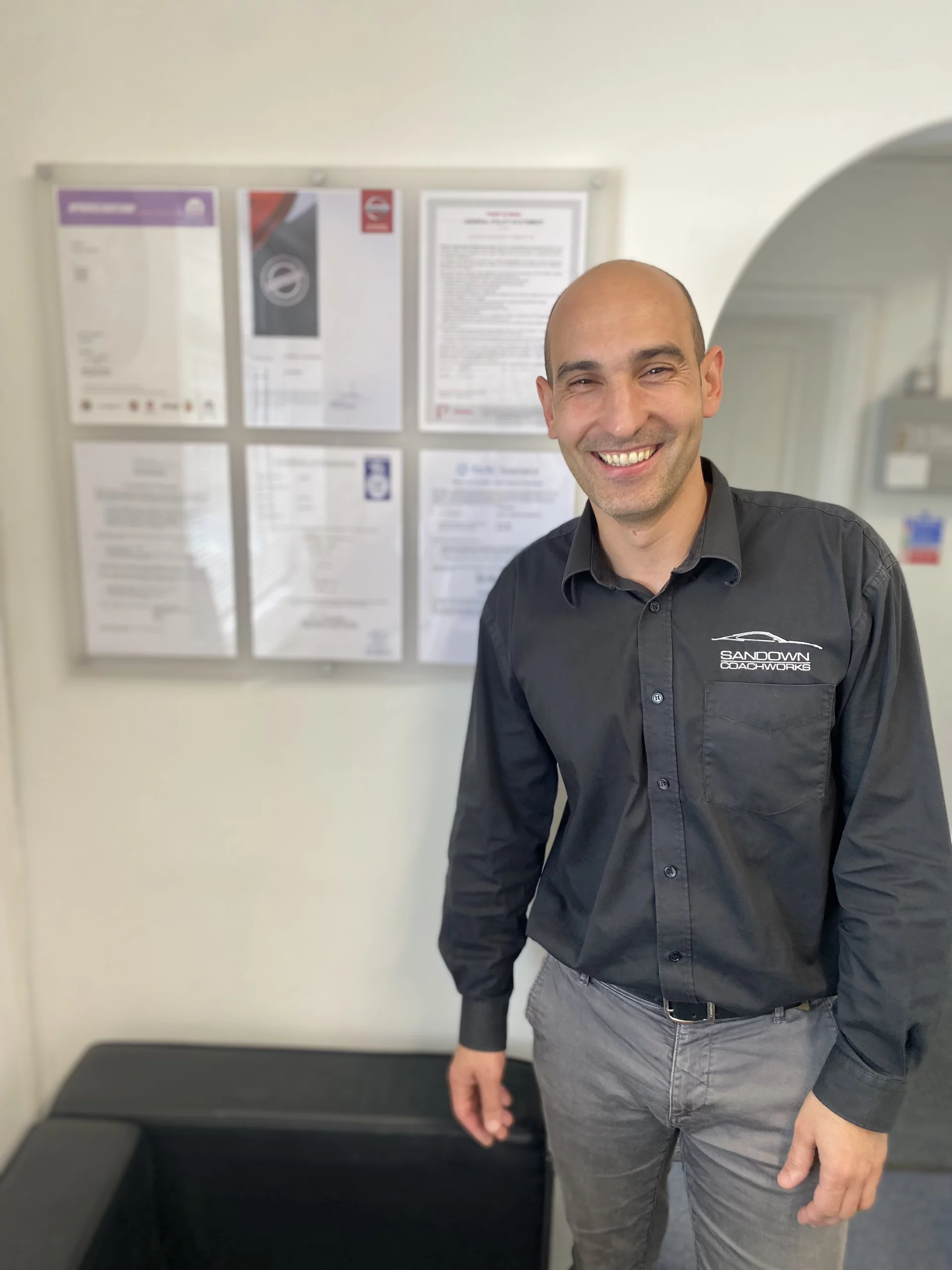 A smiling Sandown Coachworks team member standing in front of manufacturer approval certificates and BSI Kitemark accreditations.